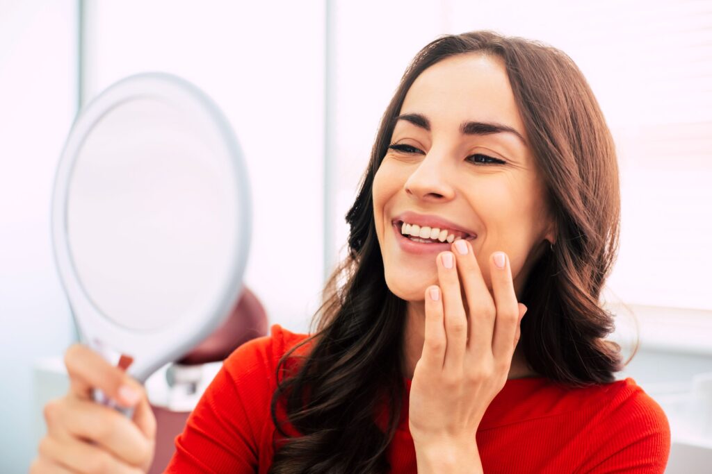 Woman with brown hair in red orange shirt smiling at reflection in handheld mirror